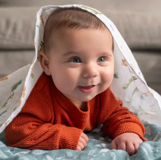 Baby in an orange sweater lying on a blanket with a patterned cover over their head. ADEN + ANAIS COTTON SWADDLE 4 PACK - DINO JUNGLE