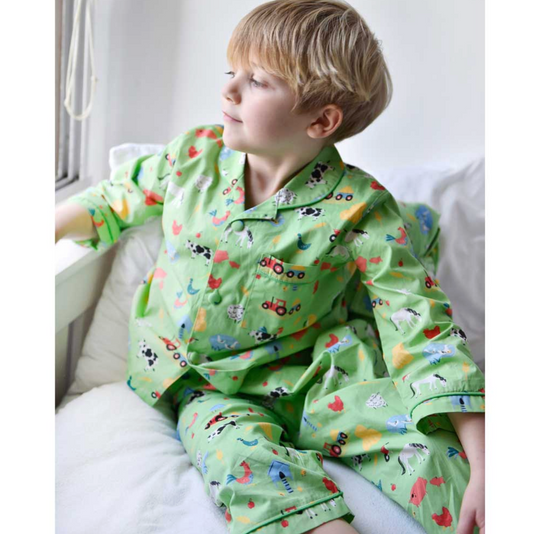 Child wearing a green pajama set with colorful patterns sitting on a white couch. CHILDREN'S CLASSIC BUTTON UP FARMYARD PYJAMAS | POWELL CRAFT