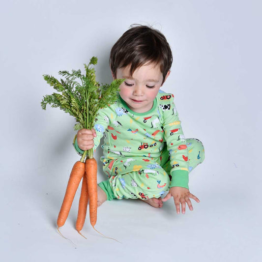 A child wearing green pyjamas with a farmyard animal print, holding a bunch of carrots with a green top. CHILDREN'S COSY FARMYARD COTTON PYJAMAS - 3 SIZES | POWELL CRAFT