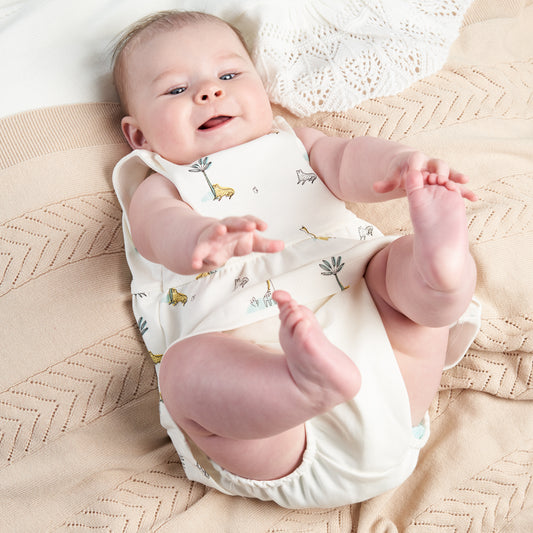 A baby lying on a textured surface wearing a white short sleeve dress with jungle animal prints and matching pants. COTTON SUMMER DRESS & PANTS - JUNGLE FRIENDS | DANDELION