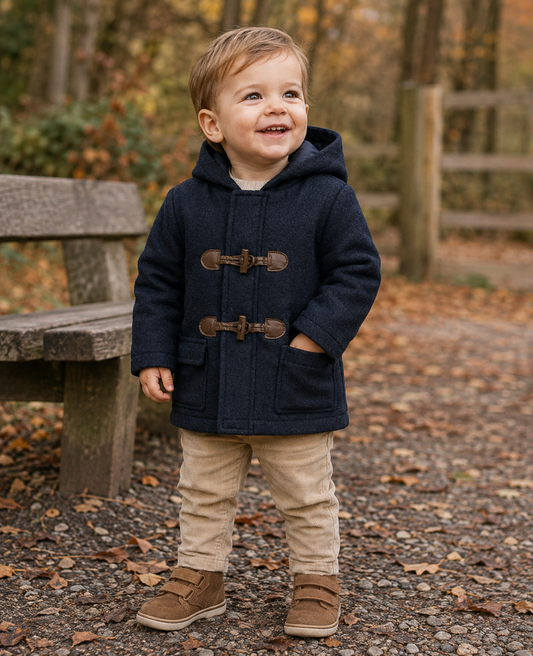 Toddler boy, approximately 18 months–2 years; gravel woodland path; autumnal golden setting; coat fully fastened, both toggles visible; hand in patch pocket; camel cords and tan boots; park bench in background.