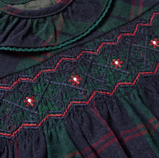 Eleanor Charles Navy & Green Tartan Smocked Dress — close-up detail of the hand-smocked chest panel; navy cable and wave smocking with red zigzag outline stitch; five red and white cross-stitch floral motifs scattered across the panel; teal-piped frill collar visible at top; gathered skirt belowCHARLES