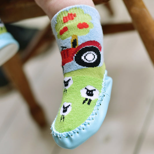 Close product view of Powell Craft pale blue tractor moccasin slipper sock; red tractor and white sheep pattern visible against a softly blurred background⠋