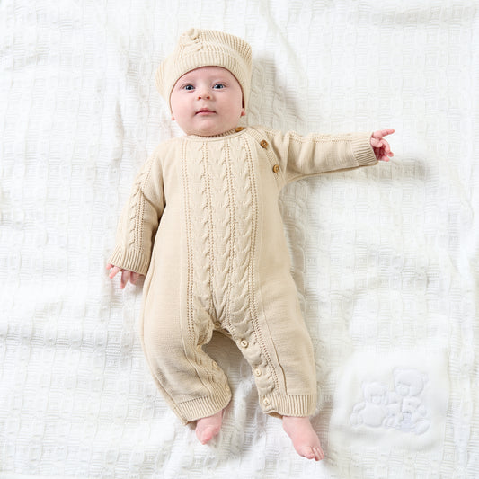 Baby lying on a white cellular blanket wearing the neutral taupe cable-knit onesie; photographed from above; central cable panel and moss-stitch side panels clearly visible; three round wooden shoulder buttons; matching cable-knit pompom hat; ribbed wrist and ankle cuffs on outstretched limbs