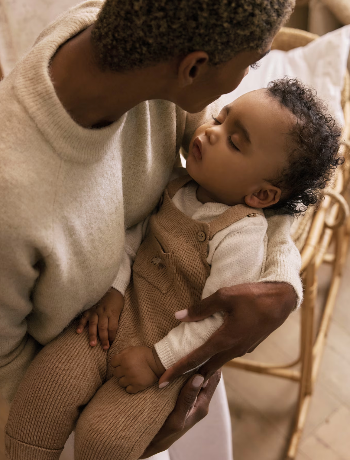 Mayoral baby 2-piece knit set in toffee — lifestyle shot of a sleeping newborn baby being cradled by a parent, wearing the toffee ribbed dungarees with wooden button bib and cream knit sweater; warm, intimate nursery setting with a rattan crib in the background