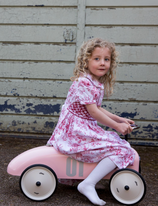 Child in a floral dress riding a pink toy car against a wooden wall. RIDE ON CLASSIC CAR - BABY PINK WITH WHITE SEAT | VILAC