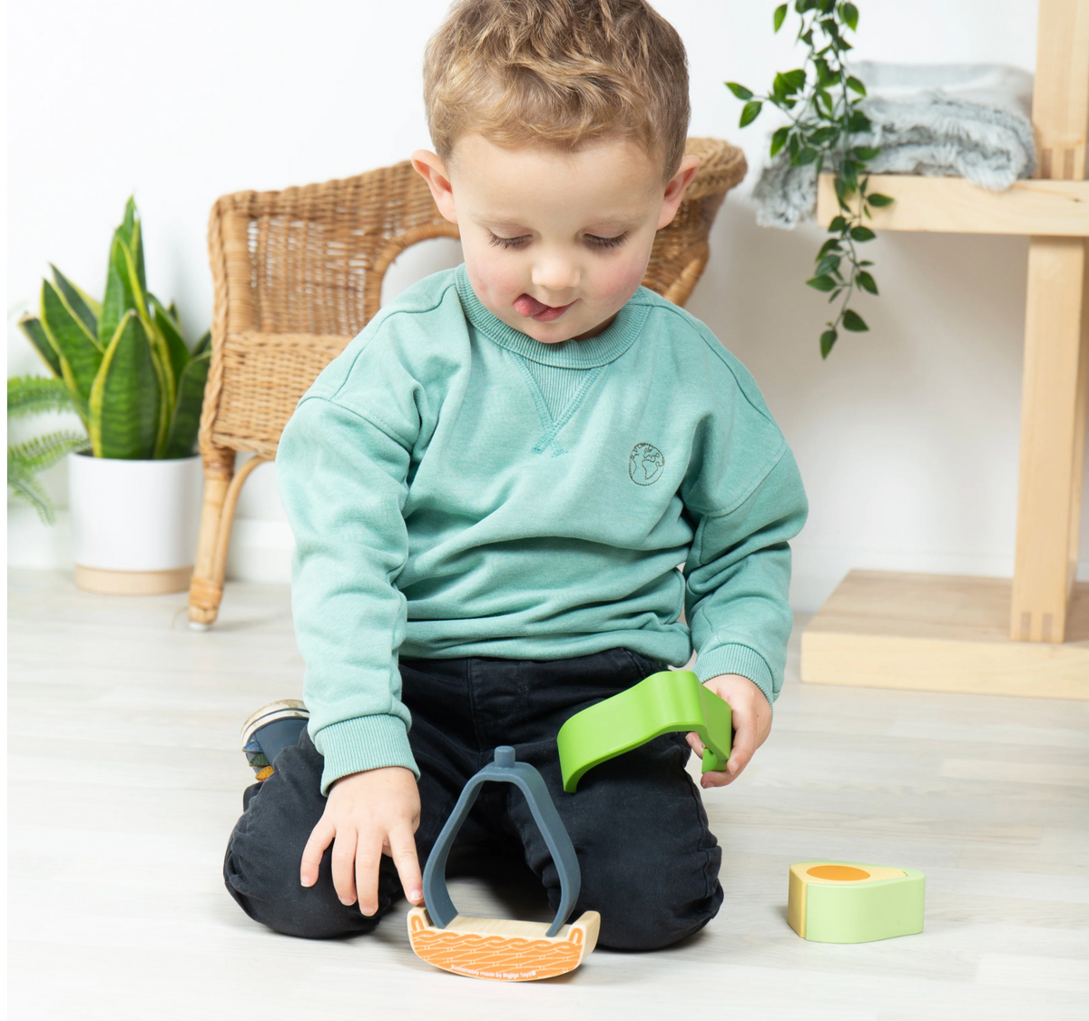 Young child seated on a light wooden floor playing with the Bigjigs Rocking Avocado Toy, handling the soft green silicone stacking pieces in a bright room with plants and pale furniture in the background.