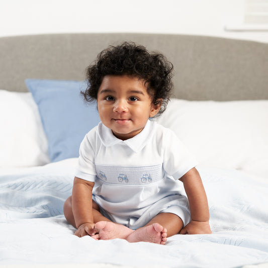 Baby in white outfit with subtle design sitting on a bed. SMOCKED TRACTOR COTTON SUMMER ROMPER OUTFIT - BABY BLUE | DANDELION