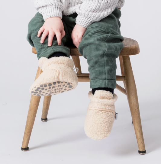 Person wearing white fuzzy slippers sitting on a wooden stool against a light background STAY-ON, NON-SLIP BOOTIE - SHERPA | THE LITTLE SOCK CO.