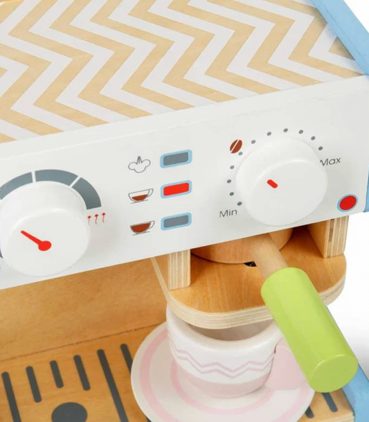 Bigjigs wooden espresso coffee maker — close-up detail of the white front panel showing the temperature gauge dial with red needle (left), printed steam, espresso and lungo cup icons with blue and red indicator lights (centre), Min/Max dial with coffee bean icon (right), and red on/off button; natural birch wood body with pale blue side panels and chevron-pattern top visible above; green portafilter handle and blush pink cup on saucer visible below on the natural wood slatted drip tray