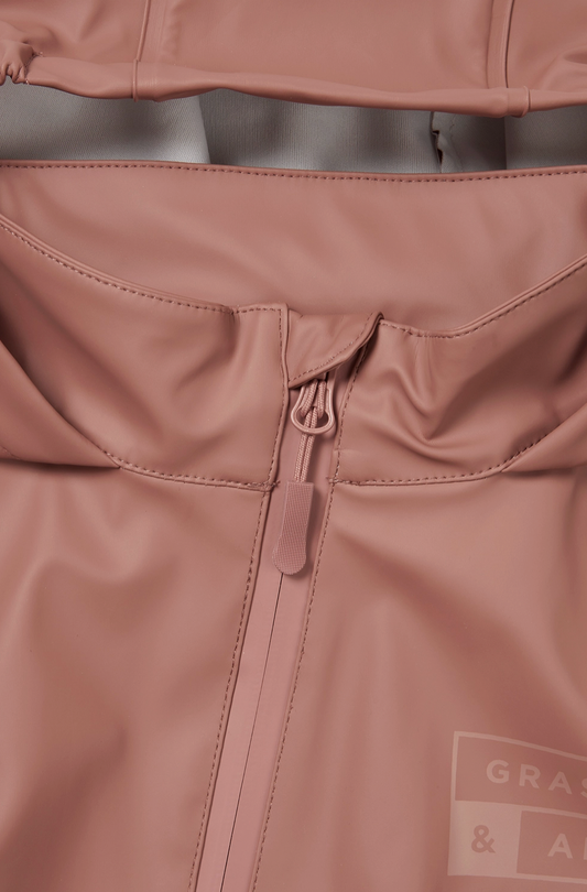 Close-up detail of the back waist panel of the Grass & Air Rose puddle suit; wide horizontal elasticated back waist panel gathered and stitched across the back of the suit creating shape and allowing a generous comfortable fit over clothes and layers; rose waterproof fabric gathered either side of the central elasticated band; taped seam stitching visible; tone-on-tone Grass & Air logo label printed in matching rose visible in the bottom right corner; zip guard flap partially visible at the top of the image