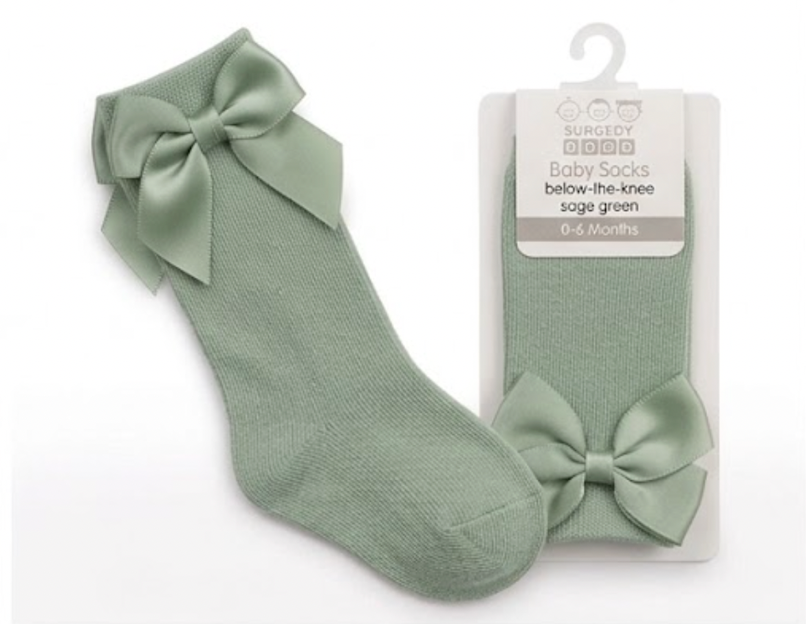 Nursery Time Baby Knee High Socks with Bow in Sage Green shown on a white background alongside original retail packaging; soft muted sage green fine-knit cotton socks with a long length sitting below the knee; each sock has a large satin ribbon bow in matching sage green at the back of the calf with two loops and two tails; retail packaging on the right is a white card hanger pack labelled Baby Socks, below-the-knee, sage green, 0 to 6 Months with the Burbery Baby brand mark
