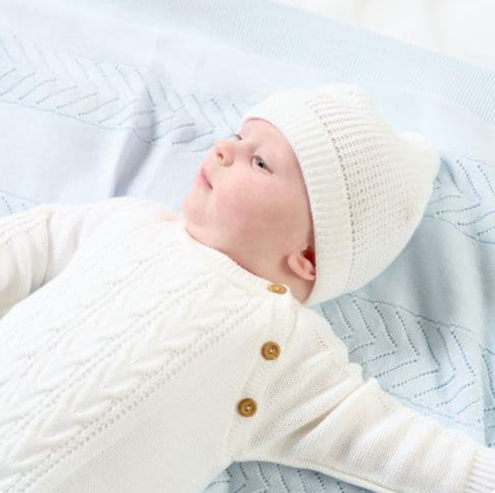 Close-up of a baby wearing the white cable-knit onesie and matching beanie; ribbed turn-up brim of the hat frames the face; cable-knit chest panel and three round wooden shoulder buttons in clear detail