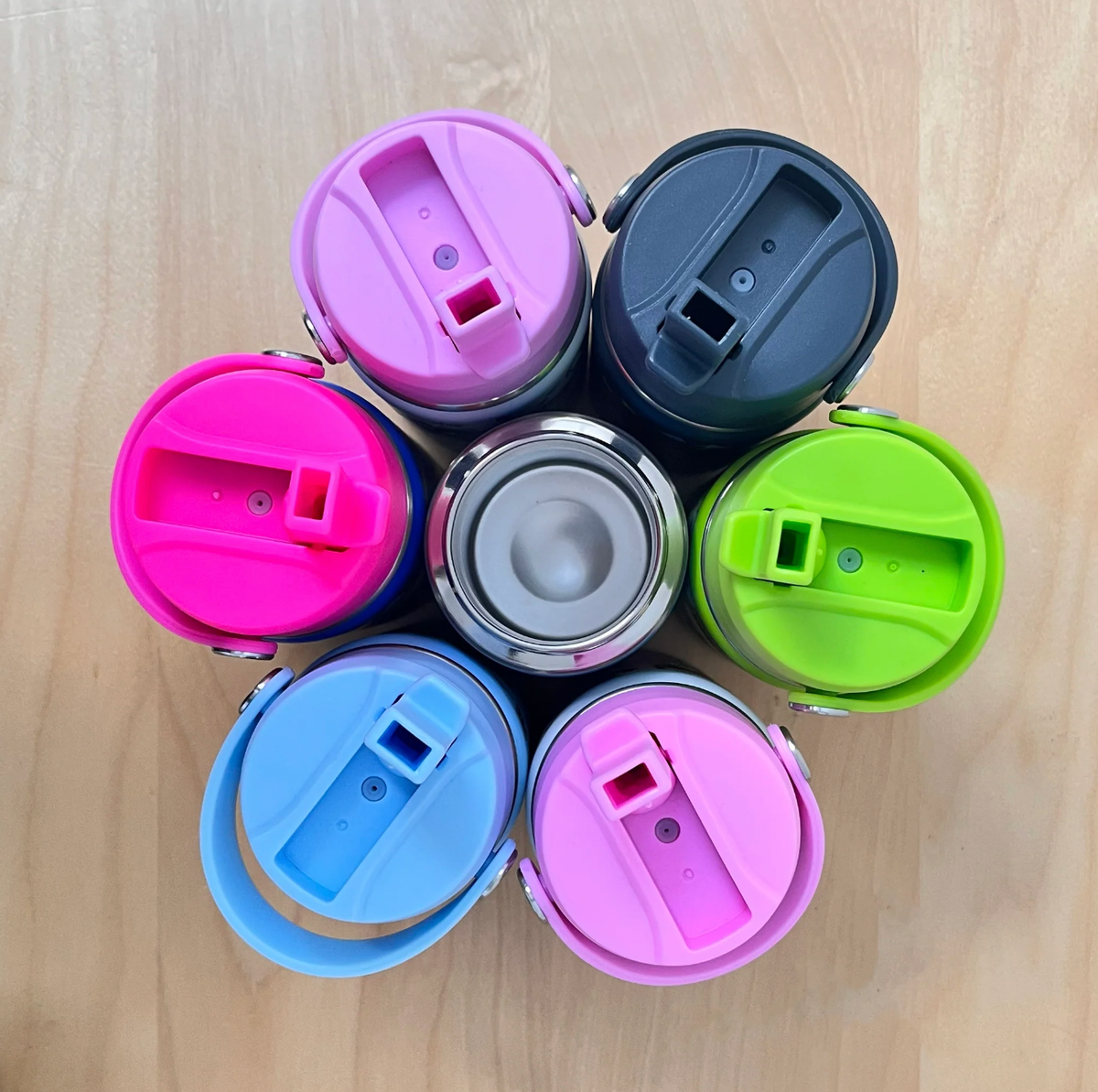 Overhead view of six Rachel Ellen straw-lid bottles encircling an open stainless steel bottle on wood; lilac and navy above, lime-green at right, pink and sky-blue below and fuchsia at left, with raised spouts and carry handles.
