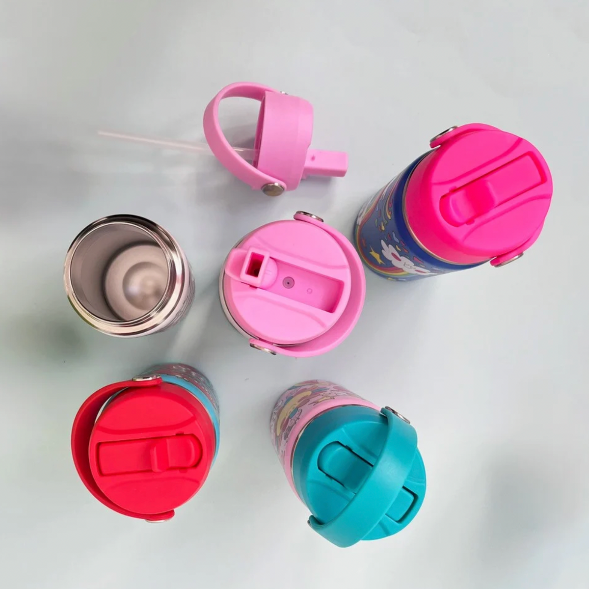 Overhead bottle range on pale blue surface showing open stainless steel bottle at left, detached pink lid with long clear straw above, raised pink spout at centre and closed fuchsia, red and turquoise lids arranged around it.
