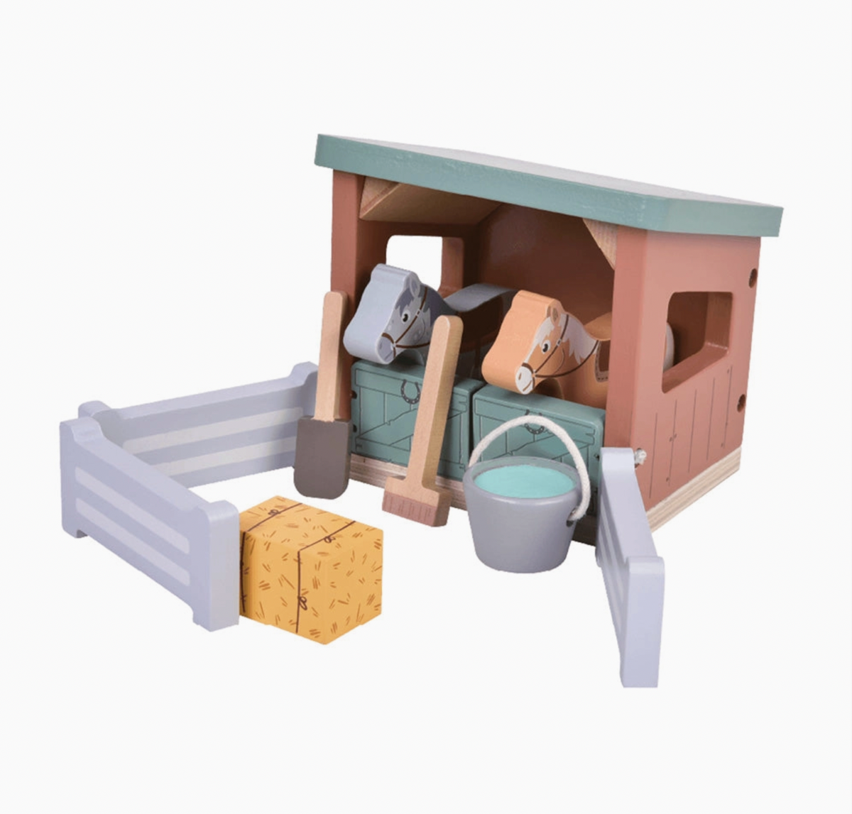 Jumini wooden horse stables set on white background: dusty-pink two-stall stable with sloping sage-green roof, matching plank-effect half doors and square side window; grey and tan horses peer out, with natural-wood-handled shovel and broom leaning in front, grey water bucket with white handle and turquoise interior, three pale-grey fences and yellow hay bale with printed straw and ties.