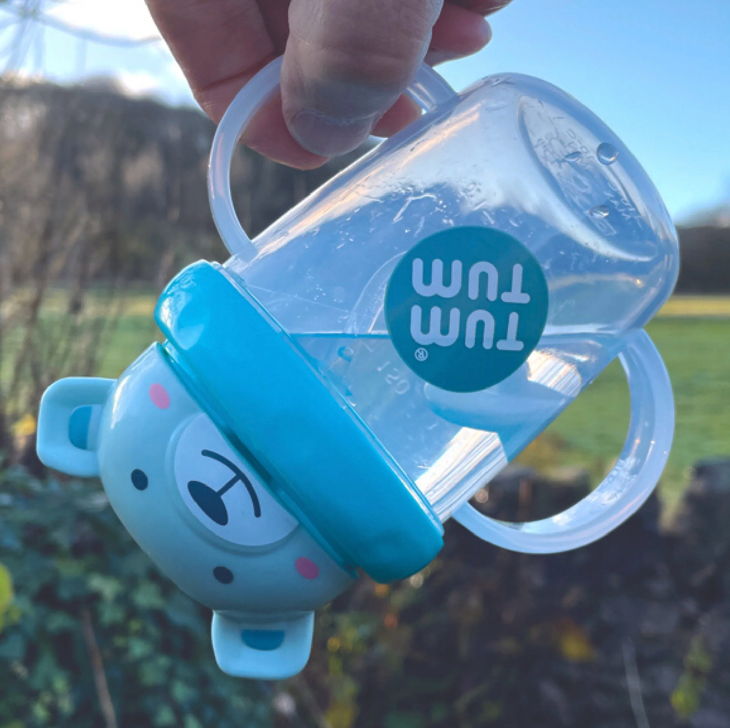 Tum Tum baby bottle with blue lid held outdoors. TIPPY CUP WITH STRAW AND FLIP TOP LEAKPROOF LID - BORIS BEAR BLUE | TUM TUM