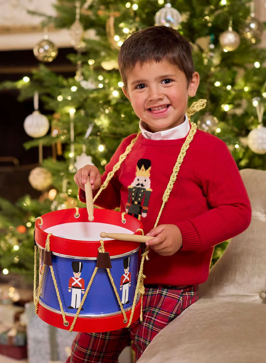 Child in a red sweater and plaid pants holding a toy drum in front of a decorated Christmas tree. TRADITIONAL TOY DRUM - THE LITTLE DRUMMER | VILAC