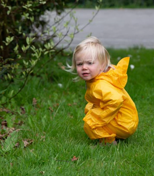 Toddler with short blonde hair aged approximately 12 to 18 months crouching low on a green grass lawn wearing the Hippychick Packasuit in sunshine yellow; hood down and windswept hair blowing; looking intently at something on the ground near a leafy green garden shrub; white Hippychick bird logo visible on the chest; grey road visible in the soft-focus background