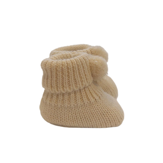 Angled side view of a beige Nursery Time knitted baby bootie pair on a white background, showing the rounded toes, ribbed cuffs, pom-poms and textured knit.
