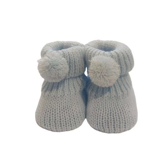 Pair of blue Nursery Time knitted baby booties on a white background, showing matching blue pom-poms, ribbed cuffs and textured knitted uppers.