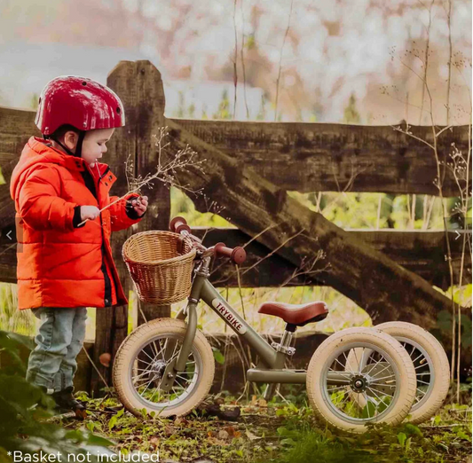 Young toddler aged approximately two to three years standing beside the Trybike Steel in vintage green trike mode in a countryside setting; child wearing a bright orange puffer jacket, jeans and a red helmet looking down at a bare winter twig in their hand; wicker basket attached to the handlebar stem (basket not included); matte olive green frame with cream tyres and chestnut brown saddle clearly visible; rustic wooden fence and early spring countryside greenery in the background
