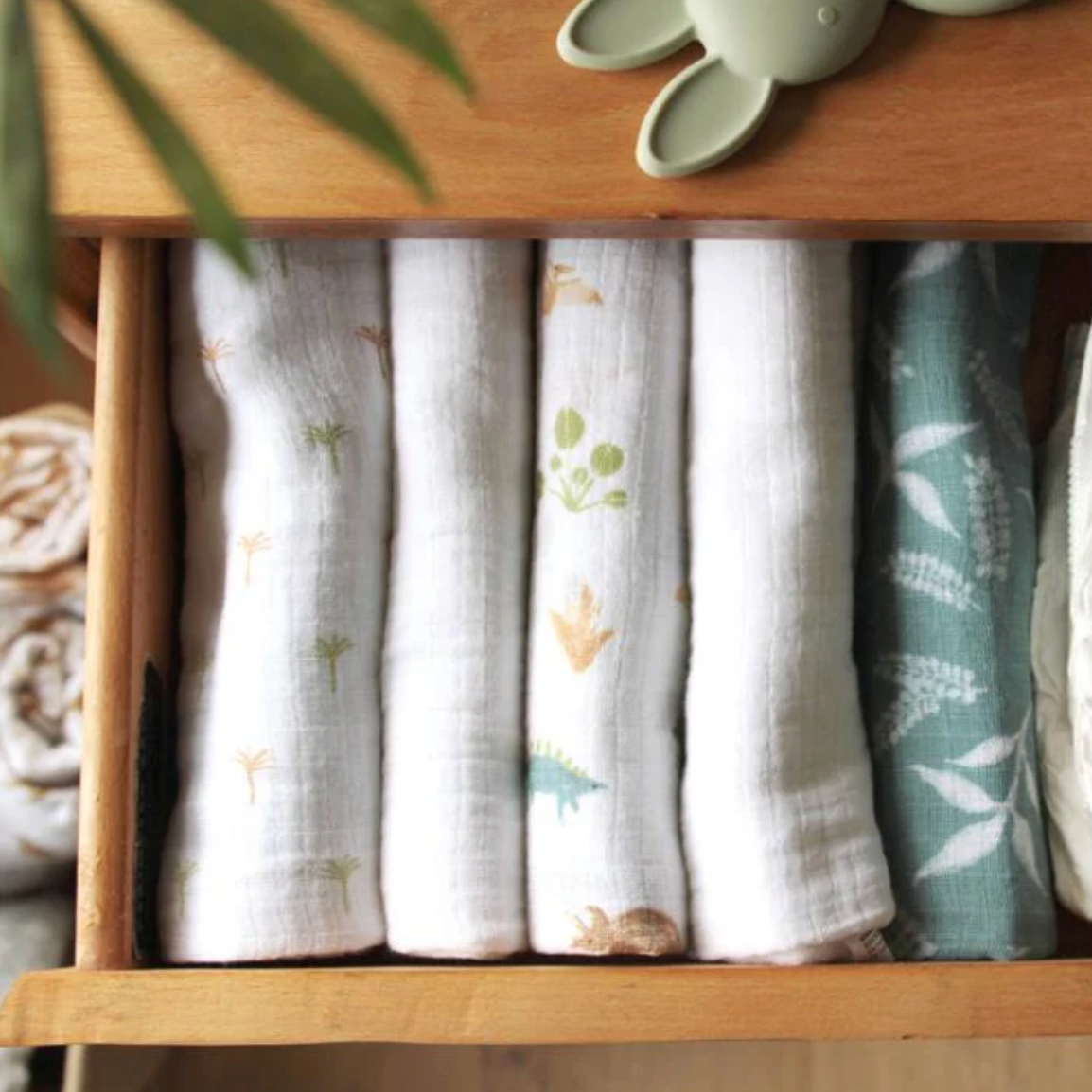 Set of folded fabric swaddles with nature-themed prints in a wooden drawer. ADEN + ANAIS 5 PACK BABY MUSLIN SQUARES - DINO JUNGLE