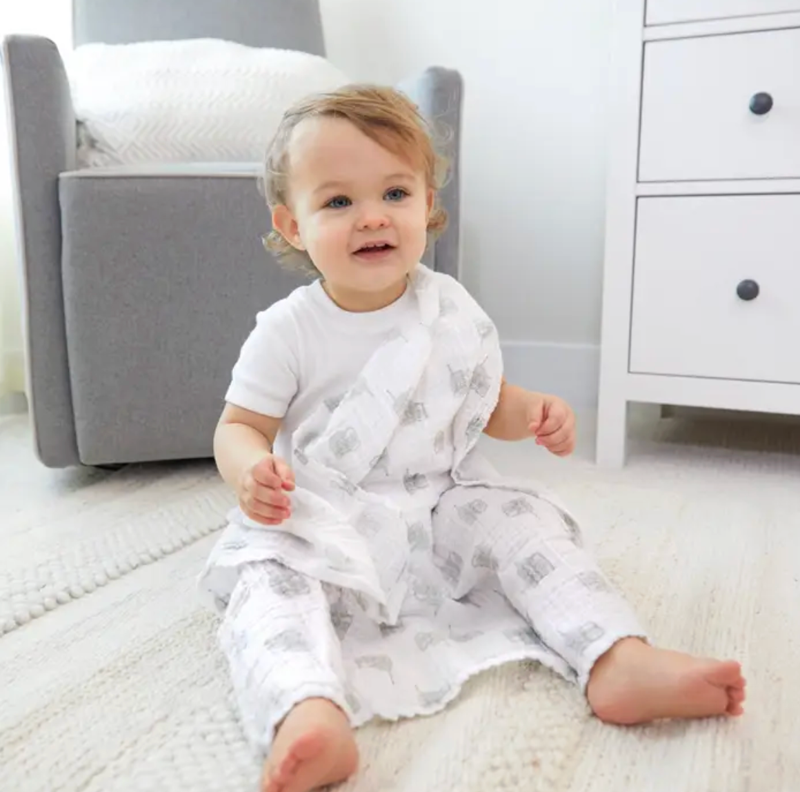 Baby sitting on a rug in a nursery with a gray chair and white dresser in the background. ADEN + ANAIS 5 PACK BABY MUSLIN SQUARES - NATURAL HISTORY