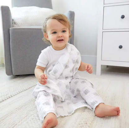 Baby sitting on a rug in a nursery with a gray chair and white dresser in the background. ADEN + ANAIS 5 PACK BABY MUSLIN SQUARES - NATURAL HISTORY