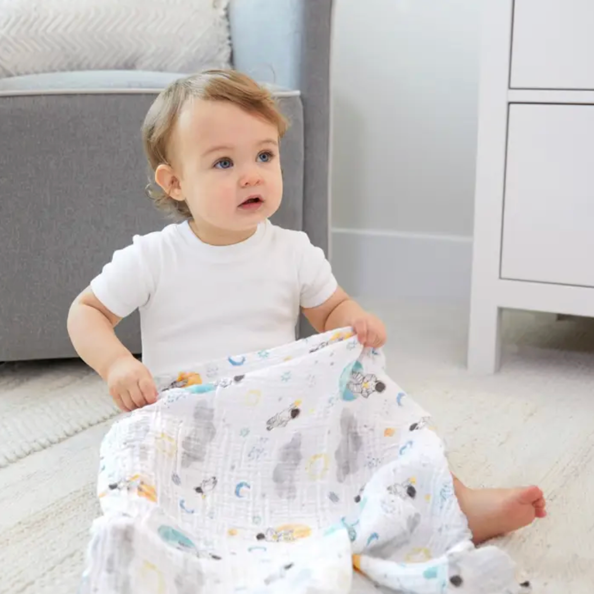 Baby sitting on a floor holding a patterned blanket in a nursery setting. ADEN + ANAIS 5 PACK BABY MUSLIN SQUARES - SPACE EXPLORERS