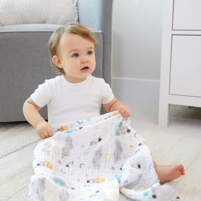 Baby sitting on a floor holding a patterned blanket in a nursery setting. ADEN + ANAIS 5 PACK BABY MUSLIN SQUARES - SPACE EXPLORERS