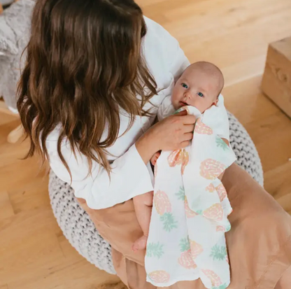 Woman holding a baby wrapped in a floral blanket on a wooden floor. ADEN + ANAIS 5 PACK BABY MUSLIN SQUARES - TROPICALIA