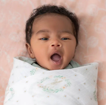 Newborn baby wrapped in a white blanket with green patterns, lying on a pink floral blanket. ADEN + ANAIS COTTON BABY SWADDLE 4 PACK - BLUSHING BUNNIES