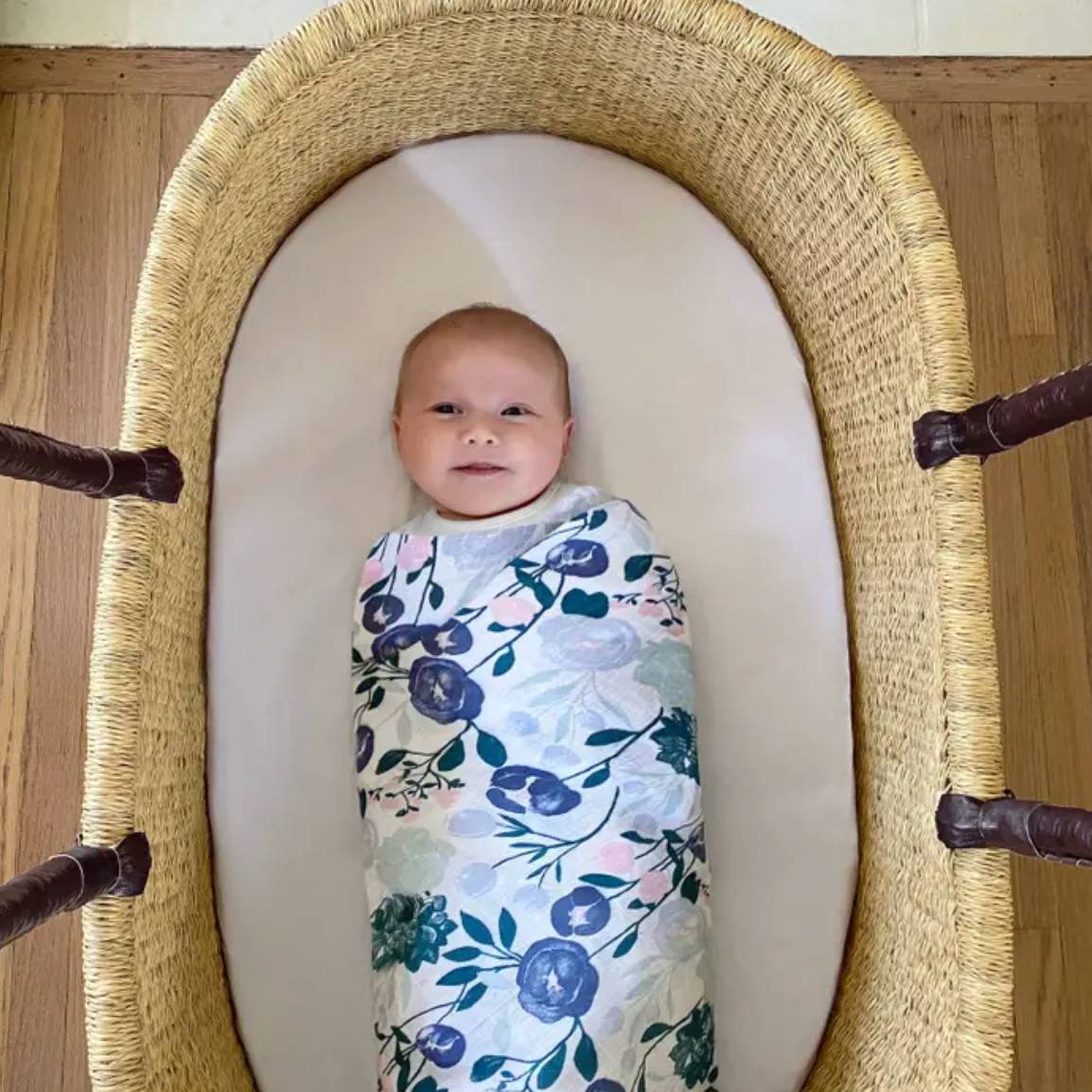 Baby swaddled in a floral blanket lying in a moses basket. ADEN + ANAIS COTTON BABY SWADDLE 4 PACK - FLOWERS BLOOM