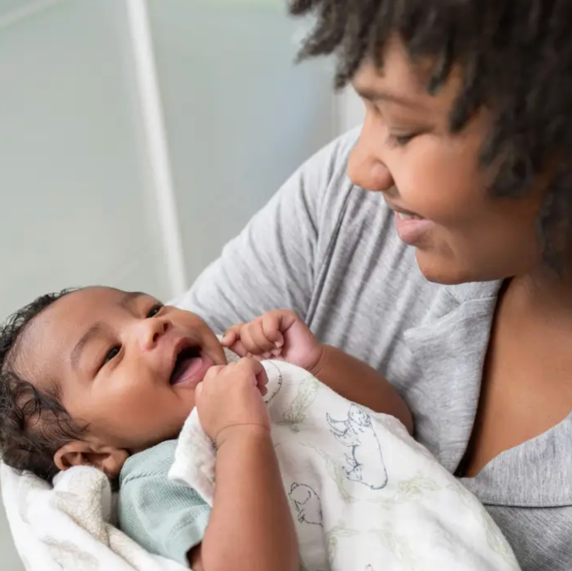 Woman holding a baby wrapped in a blanket, both looking at each other. ADEN + ANAIS COTTON BABY SWADDLE 4 PACK - HARMONY