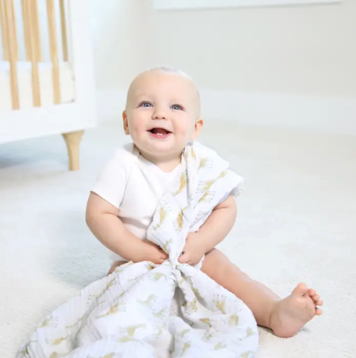 Baby sitting on a white surface with a white blanket draped over them, smiling. ADEN + ANAIS COTTON BABY SWADDLE 4 PACK - NATURAL HISTORY