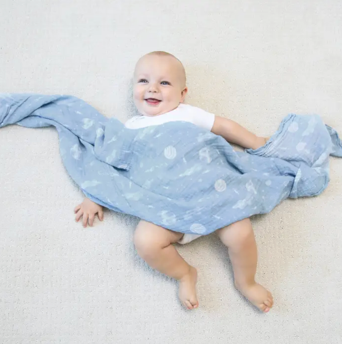 Baby lying on a light gray surface with a blue blanket featuring white patterns. ADEN + ANAIS COTTON BABY SWADDLE 4 PACK - SPACE EXPLORERS