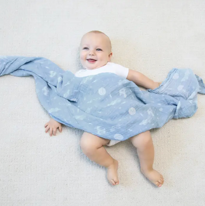 Baby lying on a light gray surface with a blue blanket featuring white patterns. ADEN + ANAIS COTTON BABY SWADDLE 4 PACK - SPACE EXPLORERS