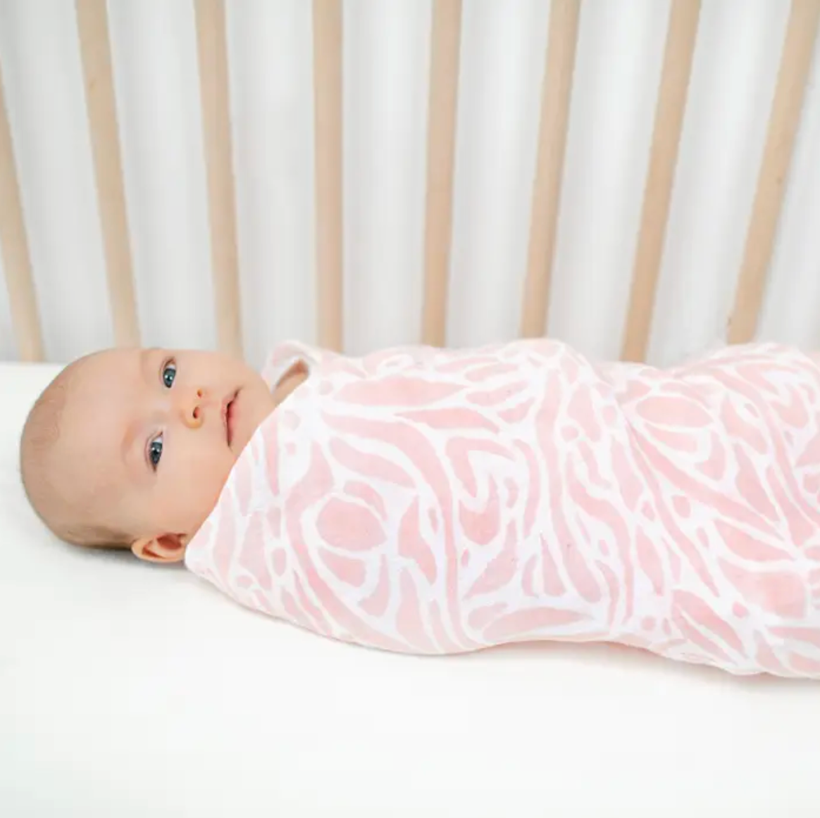 Baby swaddled in a pink floral blanket in a crib. ADEN + ANAIS COTTON BABY SWADDLE 4 PACK - TROPICALIA