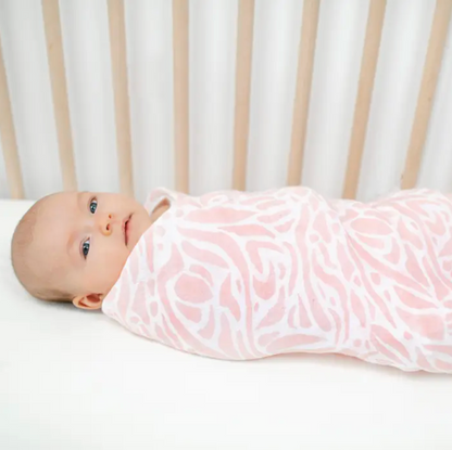 Baby swaddled in a pink floral blanket in a crib. ADEN + ANAIS COTTON BABY SWADDLE 4 PACK - TROPICALIA