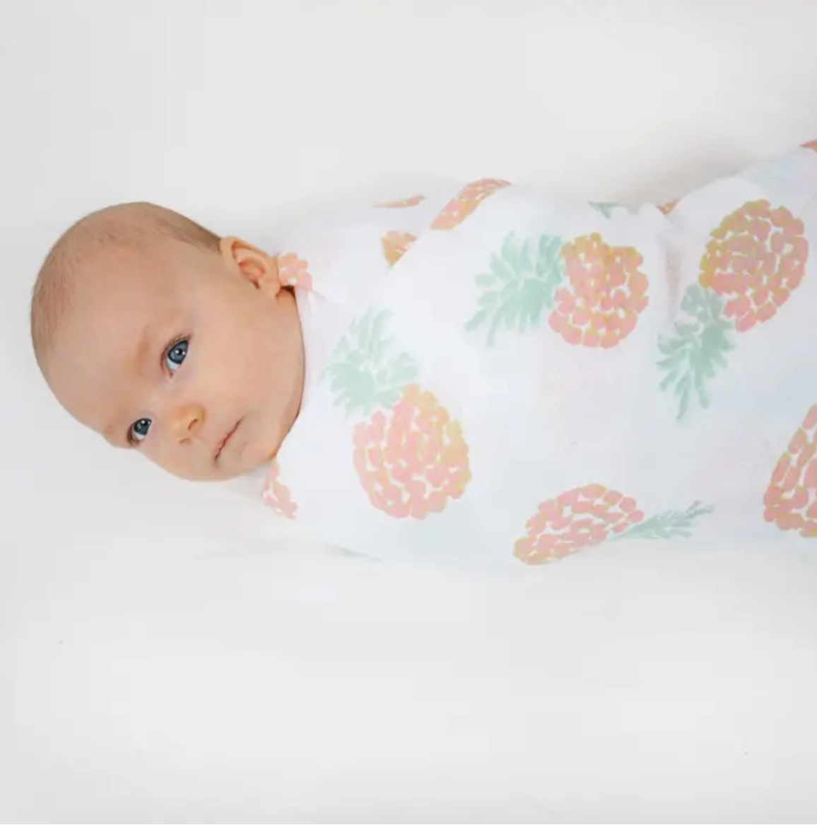 Baby wrapped in a blanket with pineapple pattern on a white background. ADEN + ANAIS COTTON BABY SWADDLE 4 PACK - TROPICALIA
