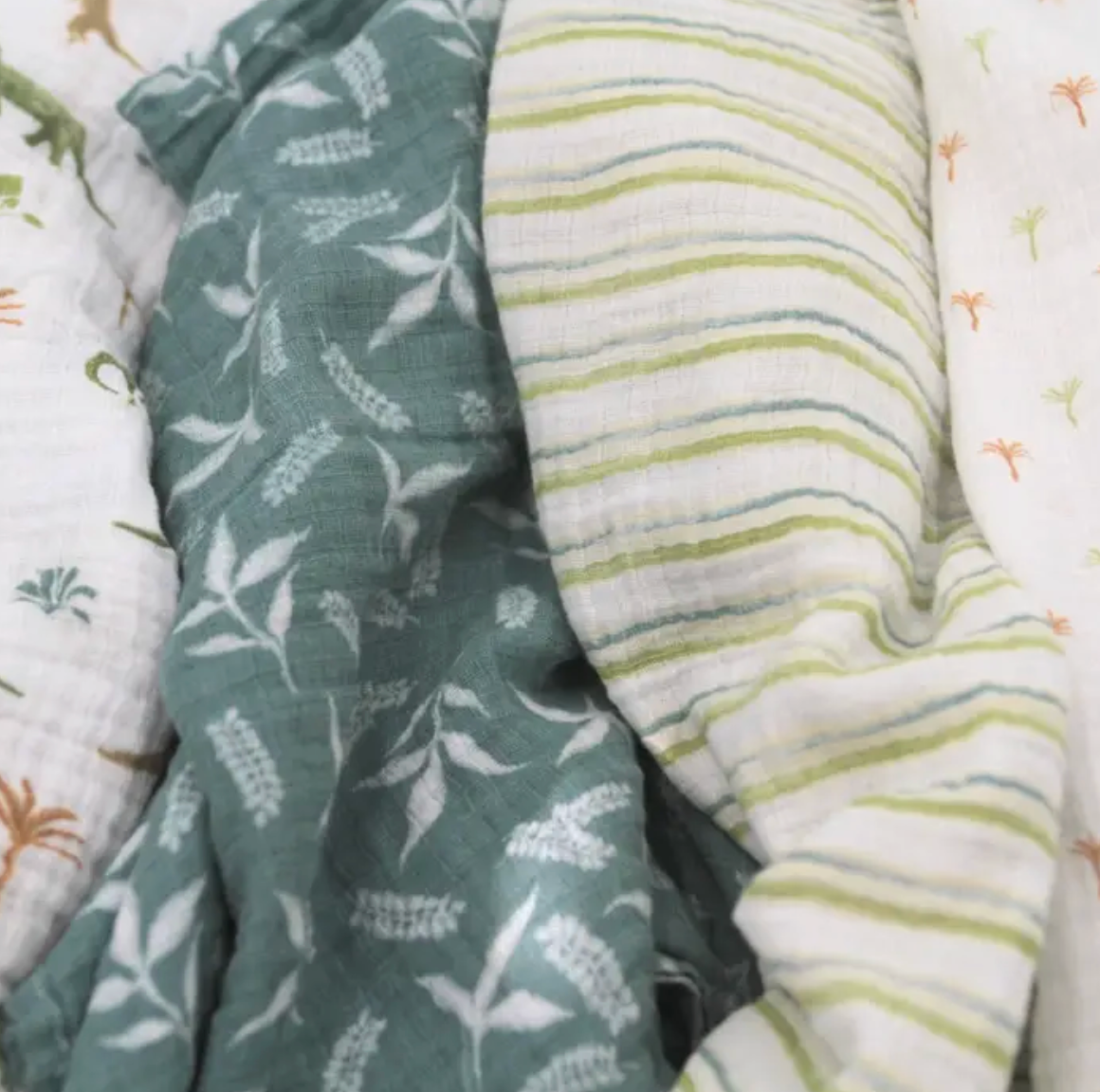 Two fabric swatches, one with a leaf pattern and the other with a striped pattern, on a textured surface. ADEN + ANAIS COTTON SWADDLE 4 PACK - DINO JUNGLE