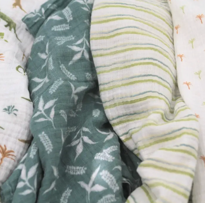 Two fabric swatches, one with a leaf pattern and the other with a striped pattern, on a textured surface. ADEN + ANAIS COTTON SWADDLE 4 PACK - DINO JUNGLE