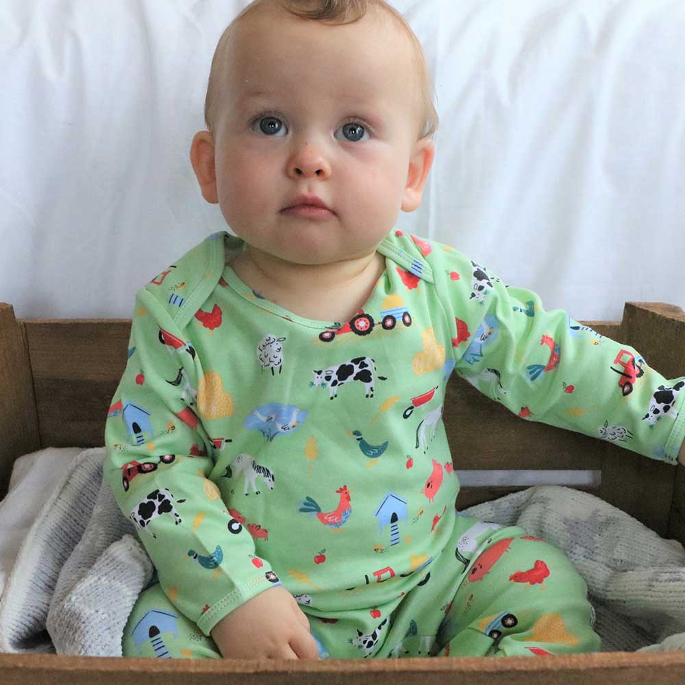 A baby wearing a long-sleeved green jumpsuit with farmyard print, including animals and farm elements. BABY COTTON FARMYARD PRINT SLEEPSUIT WITH FEET | POWELL CRAFT