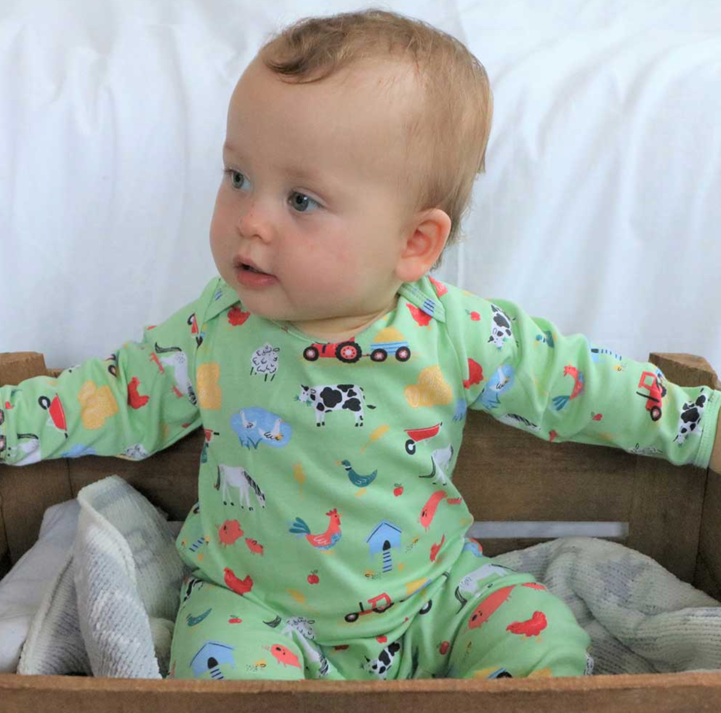 Baby wearing a green pajama with farm animal prints sitting on a wooden surface. BABY COTTON FOOTLESS FARMYARD SLEEPSUIT WITH POPPERS | POWELL CRAFT 