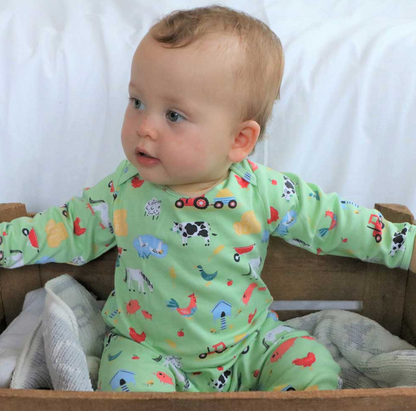 Baby wearing a green pajama with farm animal prints sitting on a wooden surface. BABY COTTON FOOTLESS FARMYARD SLEEPSUIT WITH POPPERS | POWELL CRAFT 
