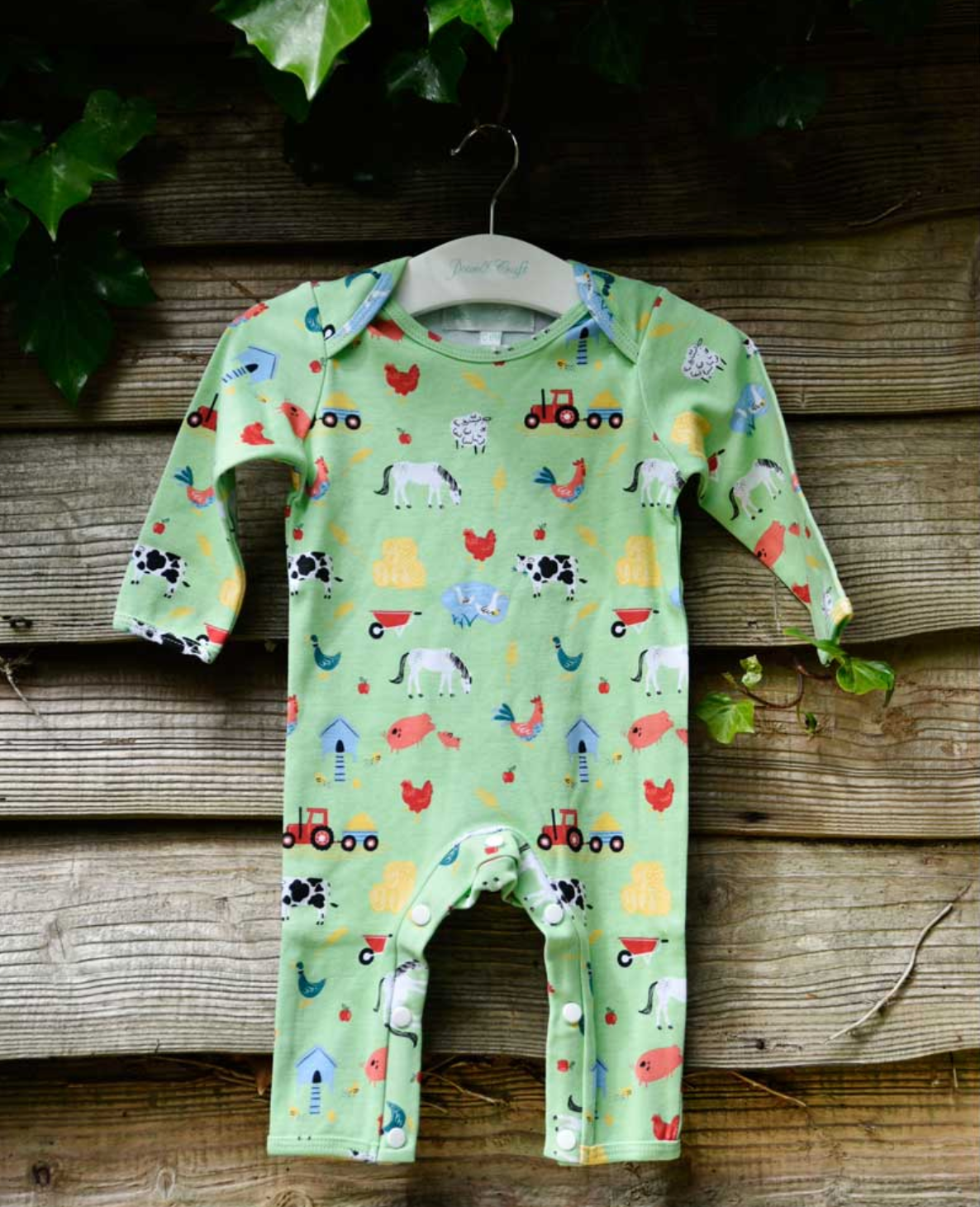 Baby romper with farm animal print on a wooden background. BABY COTTON FOOTLESS FARMYARD SLEEPSUIT WITH POPPERS | POWELL CRAFT 