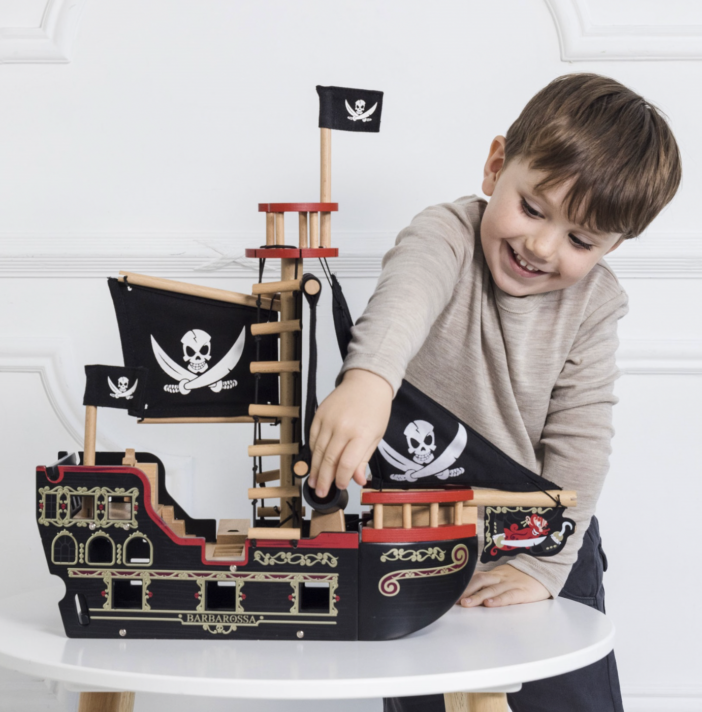 Child playing with a toy pirate ship on a white surface BARBAROSSA WOODEN PIRATE SHIP & CHARACTERS | LE TOY VAN 