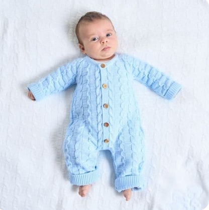 Baby boy aged approximately six to eight weeks with short dark brown hair lying on his back on a fluffy white textured blanket; wearing the Dandelion basket weave knitted onesie in soft powder blue; calm alert expression looking directly at the camera; full head-to-bare-feet view showing the complete footless onesie including the interlocking basket weave and rope cable knit texture; five warm brown buttons down the centre front; ribbed ankle cuffs at both leg ends and ribbed sleeve cuffs clearly visible; a