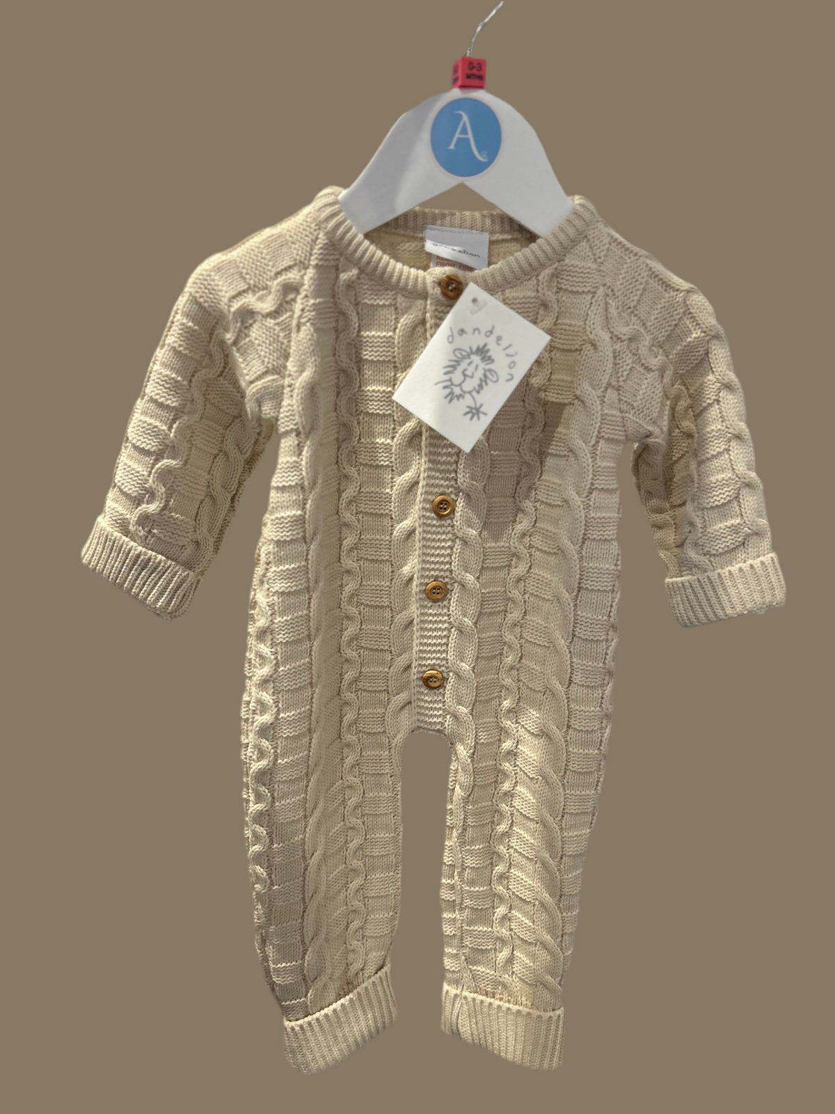 Dandelion basket weave knitted baby onesie in warm oatmeal taupe hanging on an Annabel's of Cranbrook branded white hanger with blue A roundel against a warm taupe background; Dandelion swing tag with dandelion illustration and script logo hanging from the top button; front view showing the interlocking basket weave loop panels and twisted rope cable columns of the chunky knit clearly; five round gold-toned metal shank buttons down the centre front; round ribbed neckband; ribbed cuffs visible at the sleeve 